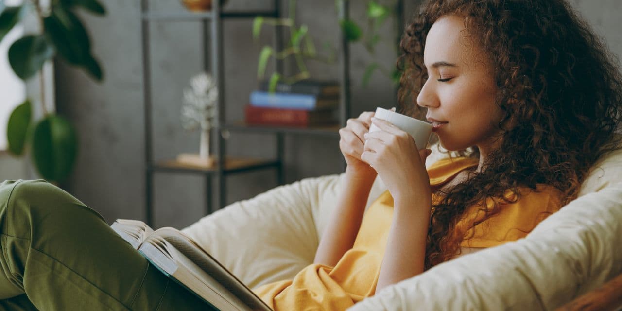 Young woman sipping on a cup of tea with her eyes closed, while sitting on a sofa with an open book on her lap.