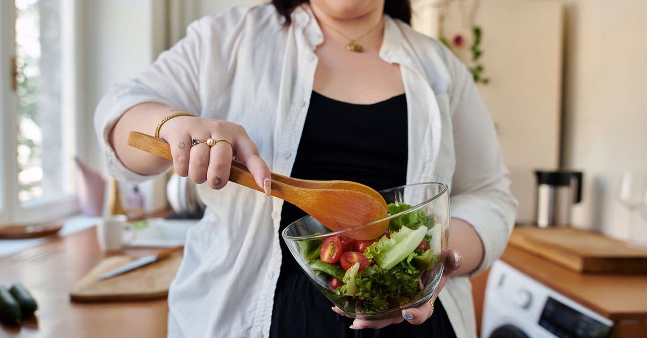 Close-up of a woman stirring a salad in a glass bowl using wooden tongs.