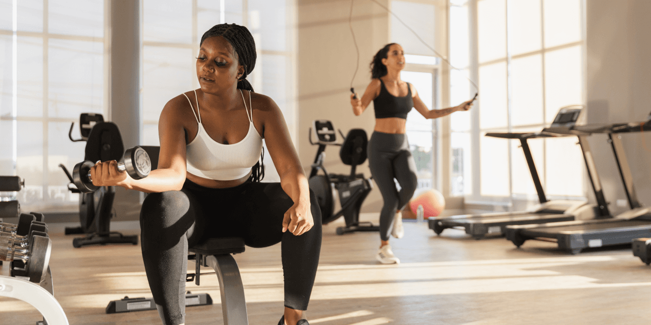 Woman lifting dumbbell in the foregroud, while another woman is skipping rope behind her in a modern gym