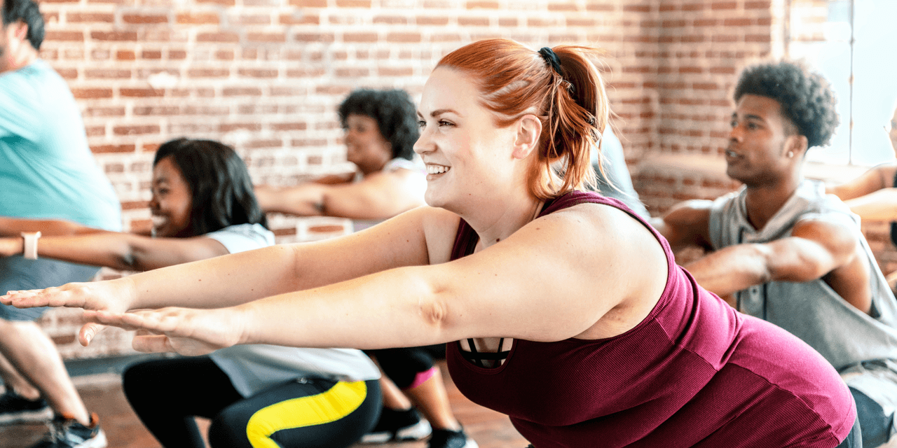 A diverse group of people having fun in a gym class, with a young woman in the foreground smiling as she performs a squat. The setting is modern and luminous, with exposed-brick walls.
